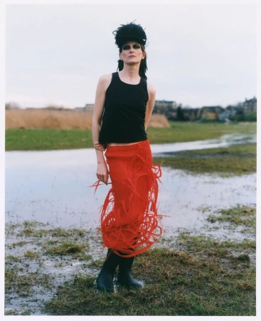 Model in a black tank top and bright red fringed skirt standing in a flooded grassy field.