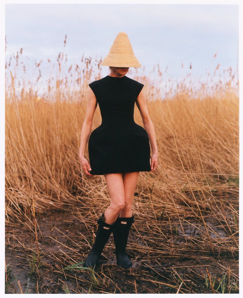 Model wearing a structured black mini dress, a tall conical straw hat covering her face, and black rain boots standing in a reed field.
