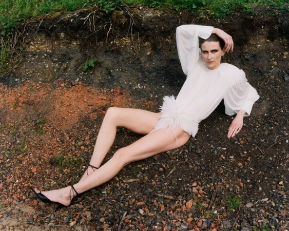 Model Arlette reclines across dark soil wearing a white long-sleeved feathered bodysuit and black strappy heels.