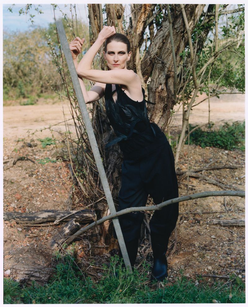 Model Arlette wearing a textured black sleeveless outfit while posing against a tree trunk in an outdoor landscape.
