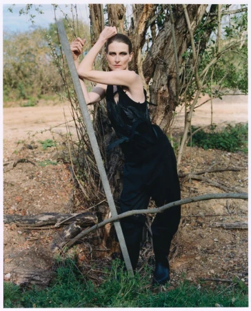 Model Arlette wearing a textured black sleeveless outfit while posing against a tree trunk in an outdoor landscape.