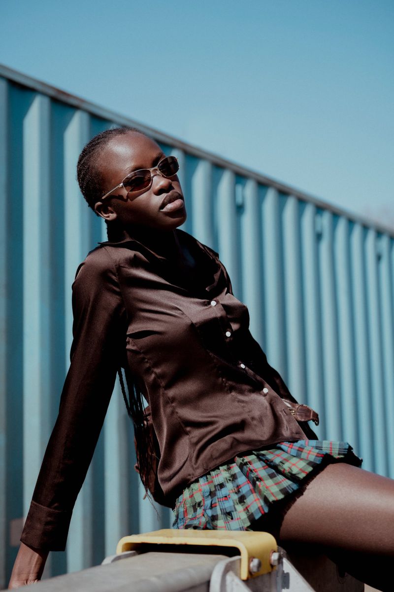 Woman in brown blouse and plaid skirt poses in outdoor medium shot with blue corrugated metal background on sunny day.