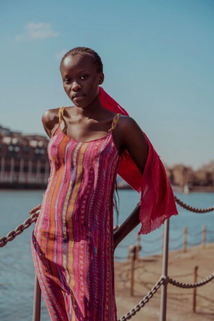 A young Black woman stands by a waterfront railing, wearing a colorful striped dress and a red scarf against a bright blue sky backdrop.