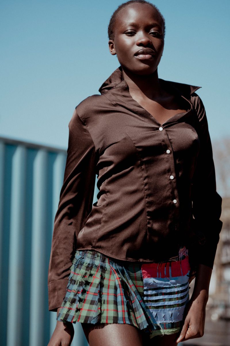 Portrait of an African woman in a satin shirt and short tartan skirt posing outside under a bright, cloudless sky.