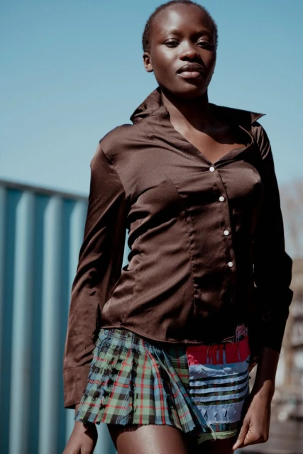Portrait of an African woman in a satin shirt and short tartan skirt posing outside under a bright, cloudless sky.
