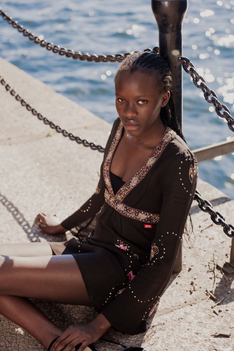 Young woman with dark hair in braids poses in a brown dress and sandals on a concrete walkway near a black metal chain railing and blue water.