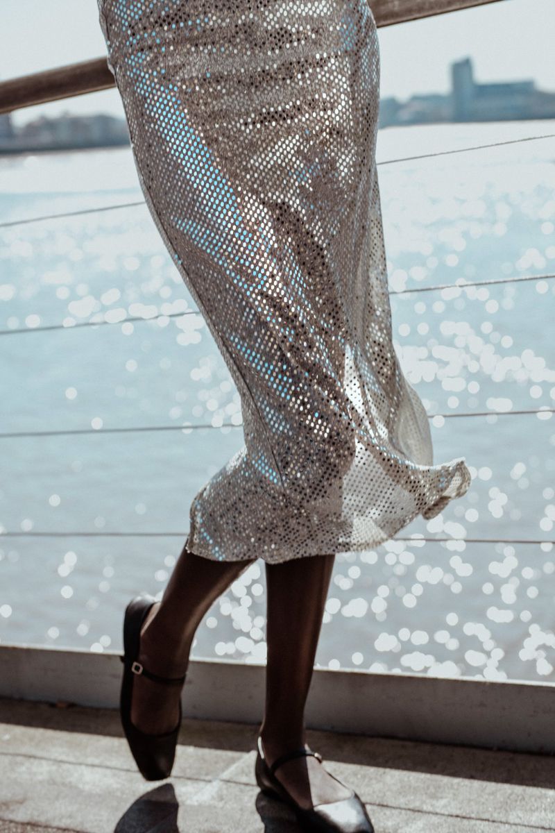 Woman in sparkling midi skirt and black Mary Jane shoes standing against harbor with city skyline in background on a sunny day.