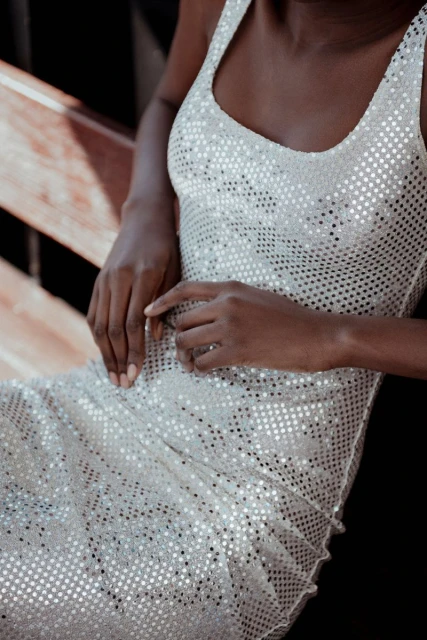 Close-up of a dark-skinned adult female wearing a sparkly silver dress, sitting on a wooden surface in an indoor setting.