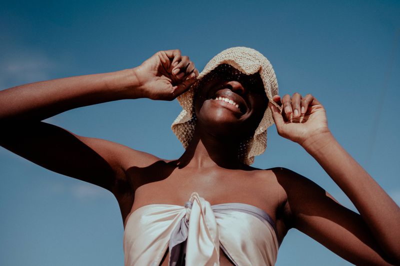 Smiling dark-skinned woman wearing a straw hat and scarf top against a clear blue sky, looking upwards with her hands near her hat.