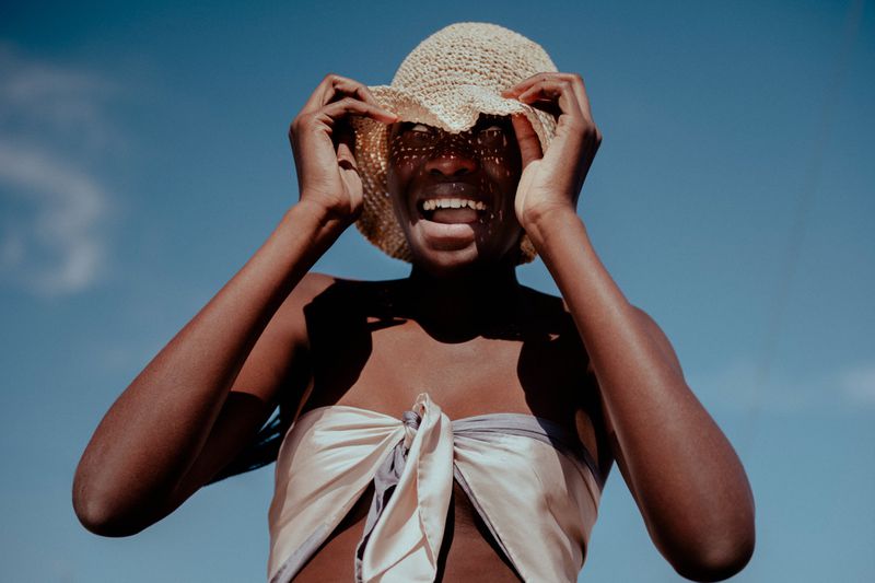 Cheerful dark-skinned woman wearing a straw hat and draped cream-colored top in a portrait against a clear blue sky on a sunny day.