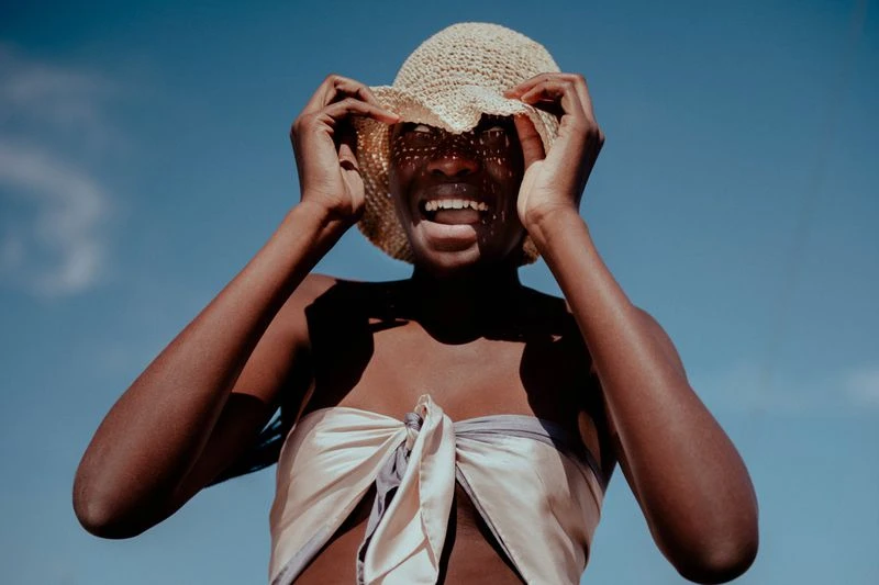 Cheerful dark-skinned woman wearing a straw hat and draped cream-colored top in a portrait against a clear blue sky on a sunny day.