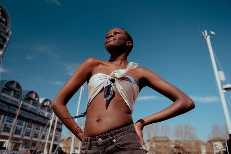 An African woman wearing a scarf top, stands against a blue sky and buildings, with her hands on her hips in the sunlight.