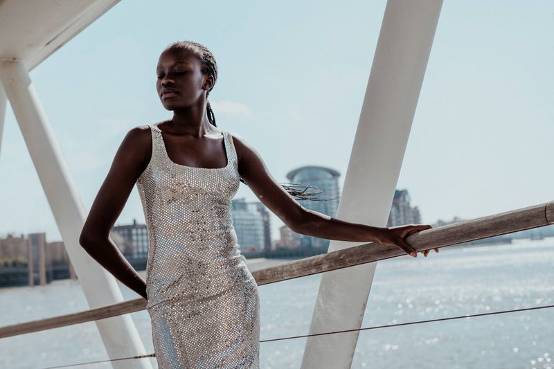 A stylish African-American woman poses outdoors in a silver sequin dress, on a bridge with a river and urban buildings in the background.