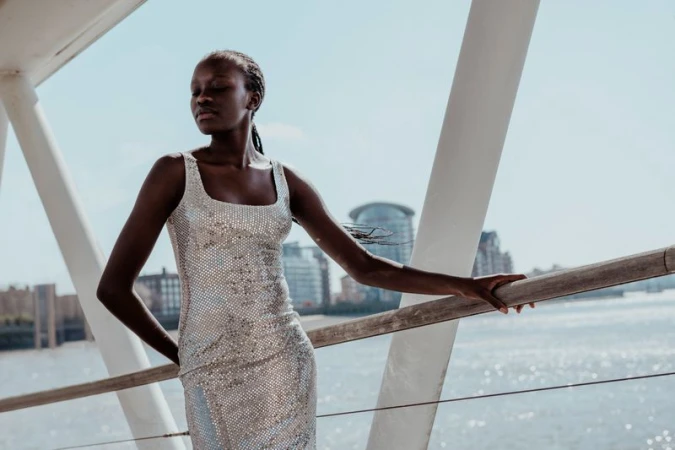 A stylish African-American woman poses outdoors in a silver sequin dress, on a bridge with a river and urban buildings in the background.