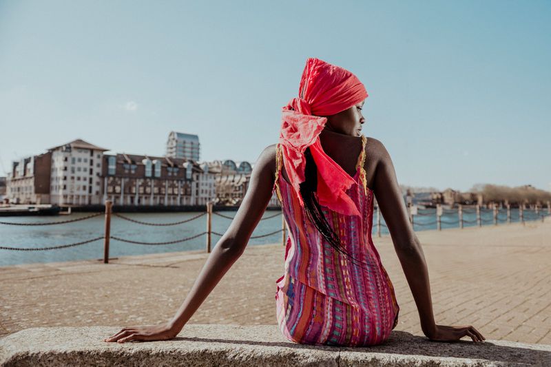 Young woman with a red headscarf and colorful dress poses in an urban setting by the water with buildings in the background on a sunny day.