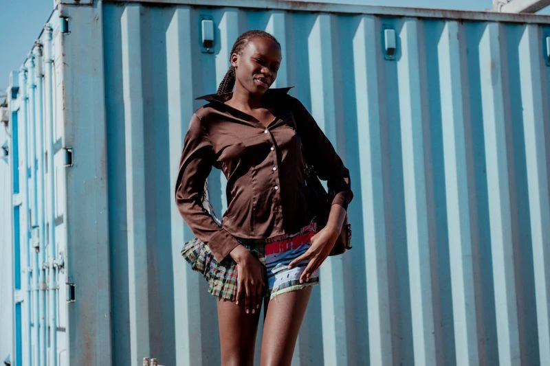 Young African woman poses outdoors wearing a brown button-down shirt and multicolored shorts in front of a corrugated metal wall.