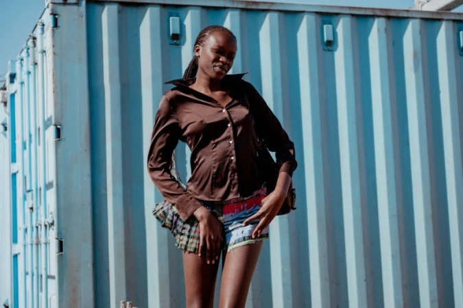 Young African woman poses outdoors wearing a brown button-down shirt and multicolored shorts in front of a corrugated metal wall.