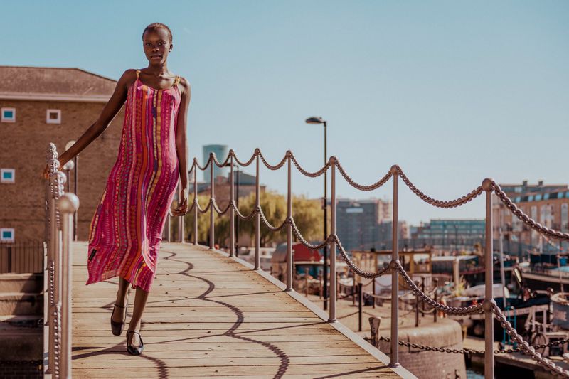 An adult female fashion model in a pink dress walks along a wooden boardwalk next to a metal chain railing with a city view backdrop.