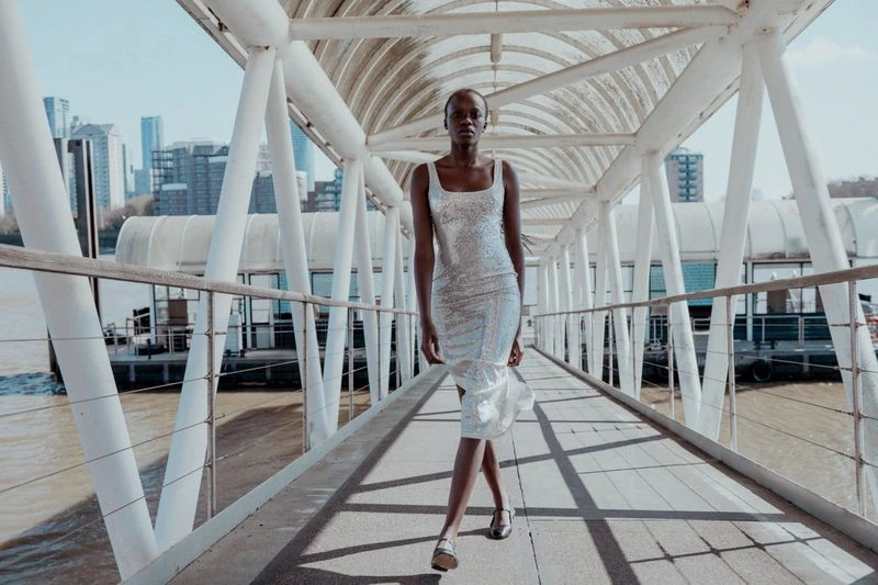 A lone Black woman walks along an elevated walkway in a sparkly dress with a city skyline and river in the background on a sunny day.