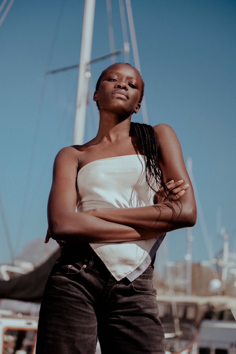 A dark-skinned woman in a white top poses with arms crossed in front of a sailboat under a clear blue sky in a portrait shot.