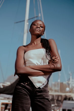 A dark-skinned woman in a white top poses with arms crossed in front of a sailboat under a clear blue sky in a portrait shot.