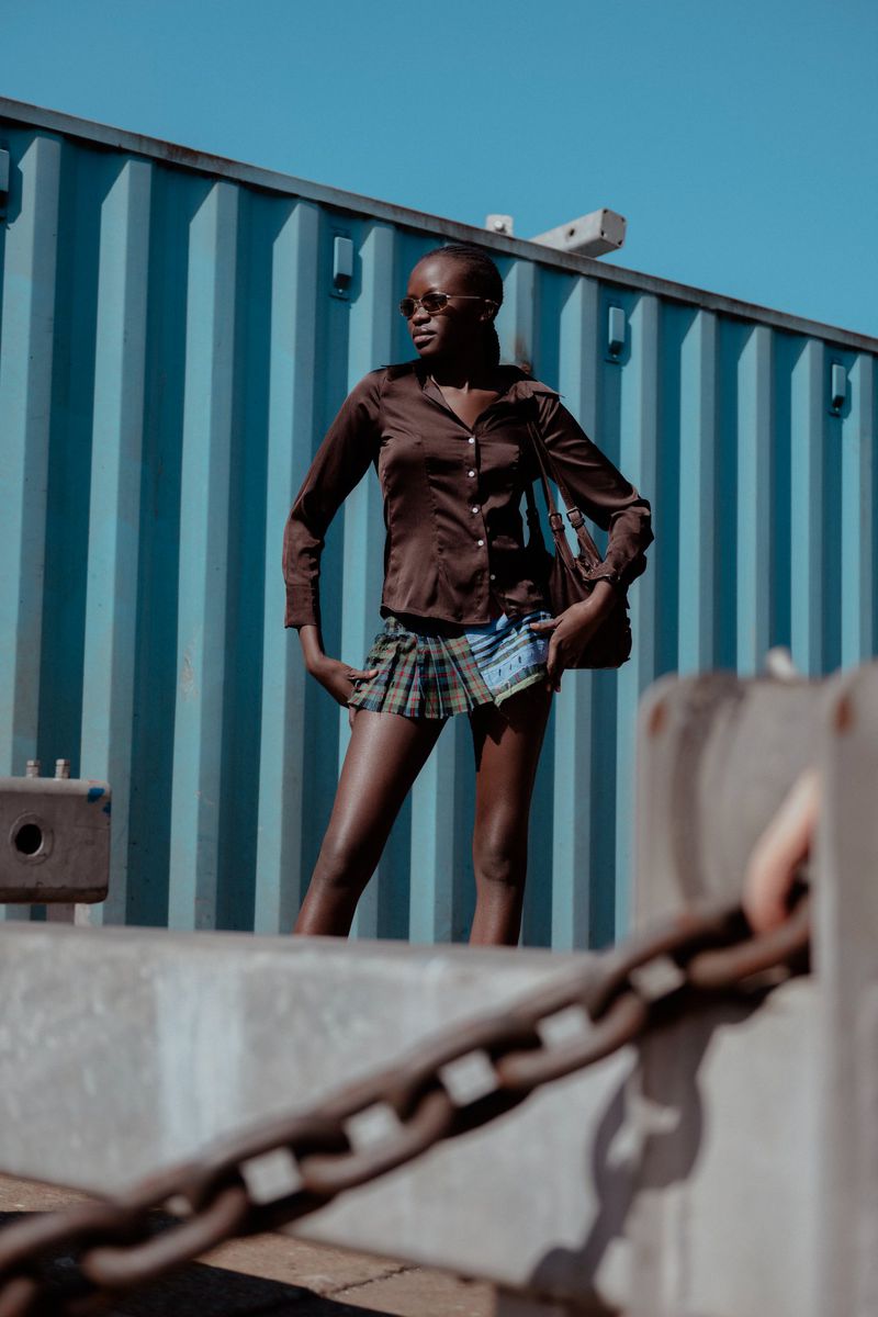 Stylish Black woman poses in brown blouse and plaid mini skirt outdoors against a blue container backdrop on a sunny day.
