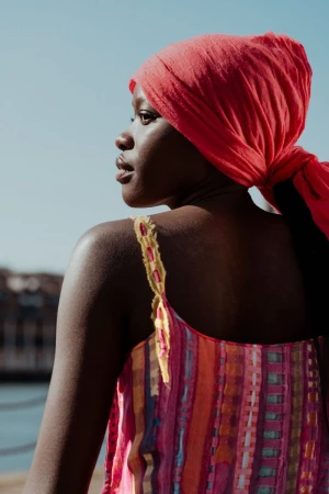 Black woman wearing a red headscarf and a colorful patterned strap top against a clear sky