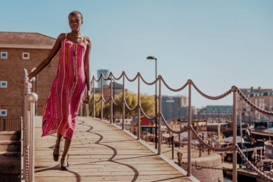 Black woman wearing a colourful patterned slip dress walking on a wooden pier with chain railings.