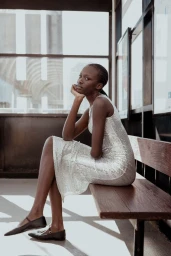 Black model in a sequin dress seated on a wooden bench inside a transit shelter