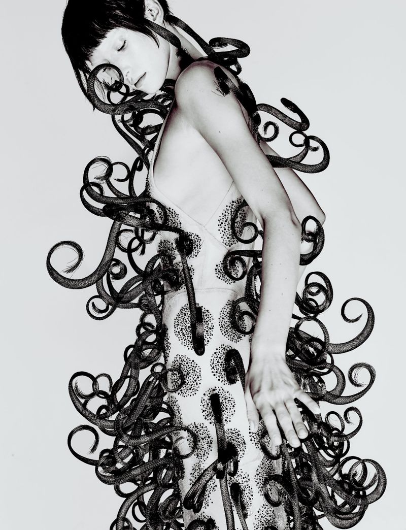 Model posing in an embellished pale slip dress intertwined with swirling black sculptural tendrils against a white studio backdrop.