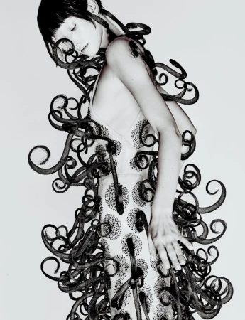 Model posing in an embellished pale slip dress intertwined with swirling black sculptural tendrils against a white studio backdrop.