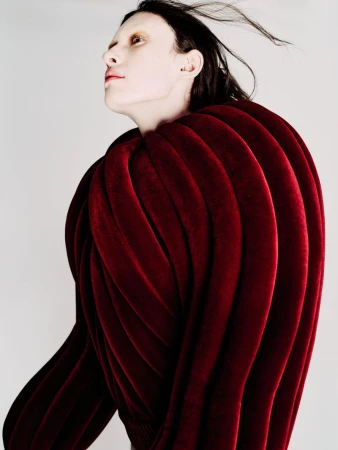 Model wearing a sculptural ribbed deep red velvet top against a plain light background, looking upward with windblown hair.