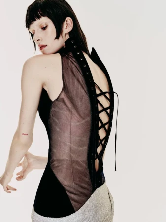 Model facing away in a black sleeveless lace-up corset top with a sheer back panel against a white backdrop.