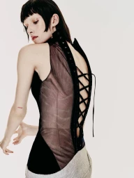 Model with dark bangs posing in profile wearing a black sleeveless corset top with a lace-up sheer back against a white background.