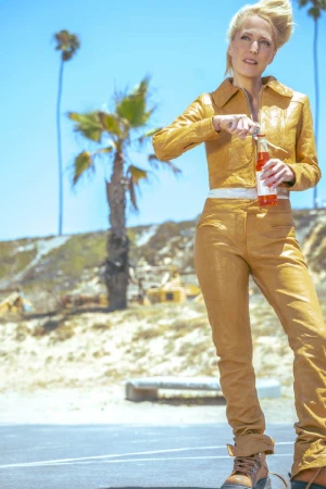 Gillian Anderson standing outdoors in a mustard yellow leather jacket and pants opening a glass bottle under palm trees.