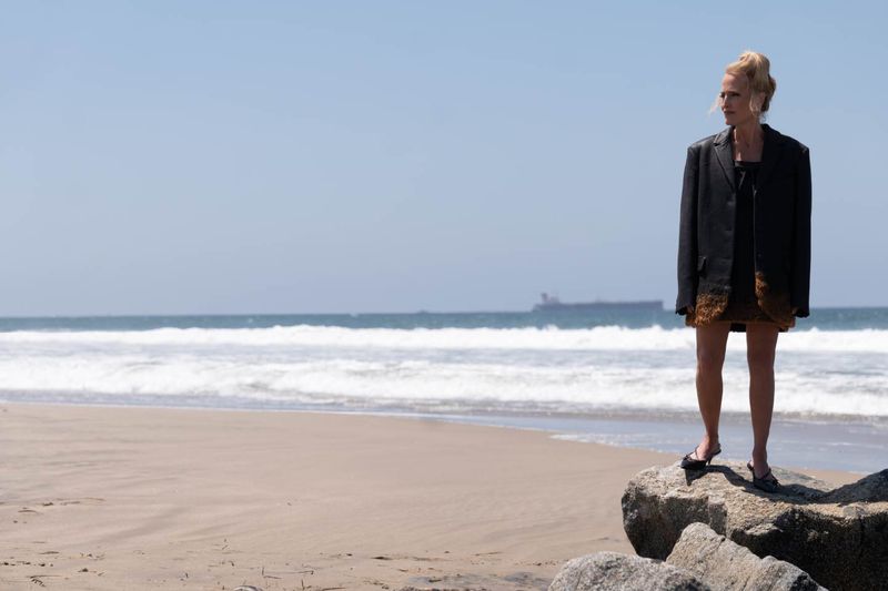 Gillian Anderson in an oversized black blazer and heels standing on coastal rocks at a sandy beach.