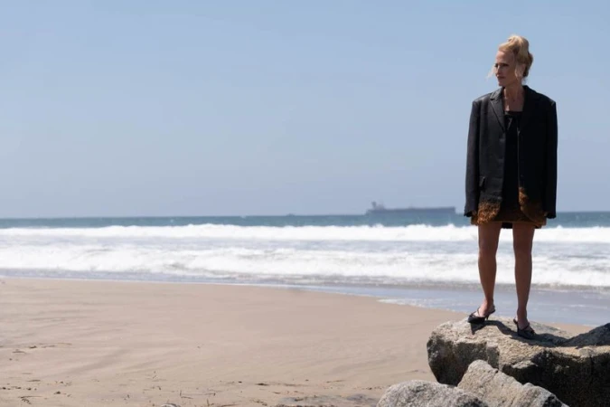 Gillian Anderson in an oversized black blazer and heels standing on coastal rocks at a sandy beach.