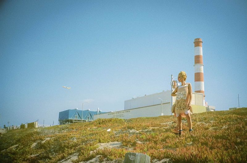 Gillian Anderson poses on a grassy hillside before an industrial facility and red-and-white smokestack under a clear sky.