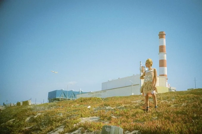 Gillian Anderson poses on a grassy hillside before an industrial facility and red-and-white smokestack under a clear sky.