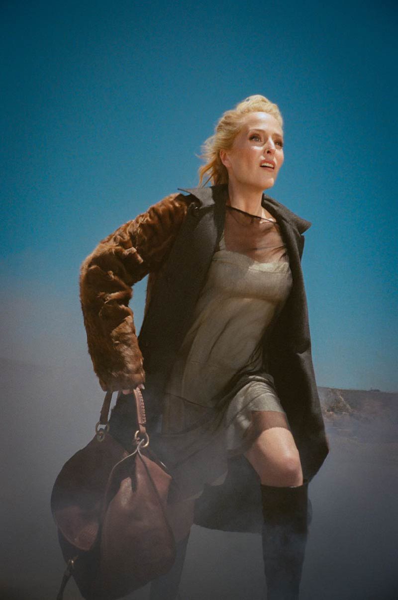 Gillian Anderson walks forward in a black coat with brown fur sleeves, slip dress, and carrying a leather handbag against a blue sky.