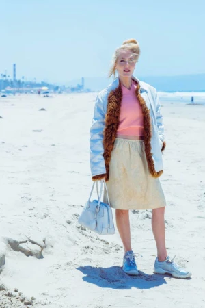 Gillian Anderson stands on a sandy beach wearing a fur-lined coat, pink top, beige skirt, sneakers, and holding a handbag.
