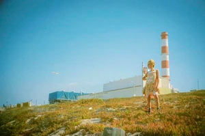 Gillian Anderson standing outdoors on a grassy hillside with an industrial building and red-and-white smokestack behind her.