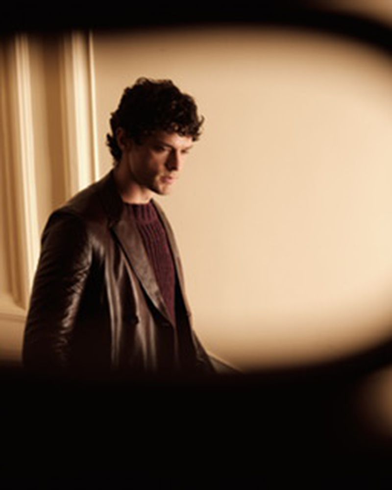 Model Marin Fabre in a dark brown leather coat and ribbed sweater, seen through a dark foreground aperture.