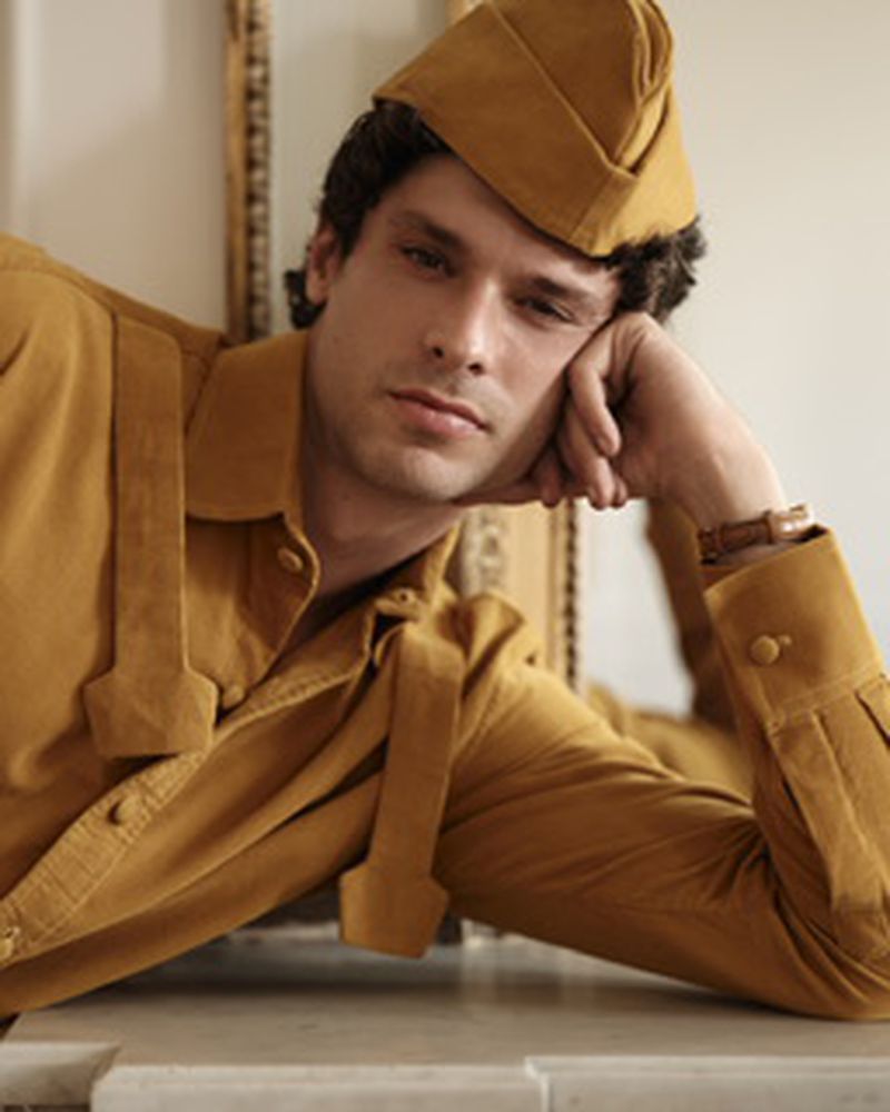 Model Marin Fabre resting his chin on his hand wearing a mustard yellow buttoned shirt and matching garrison cap.