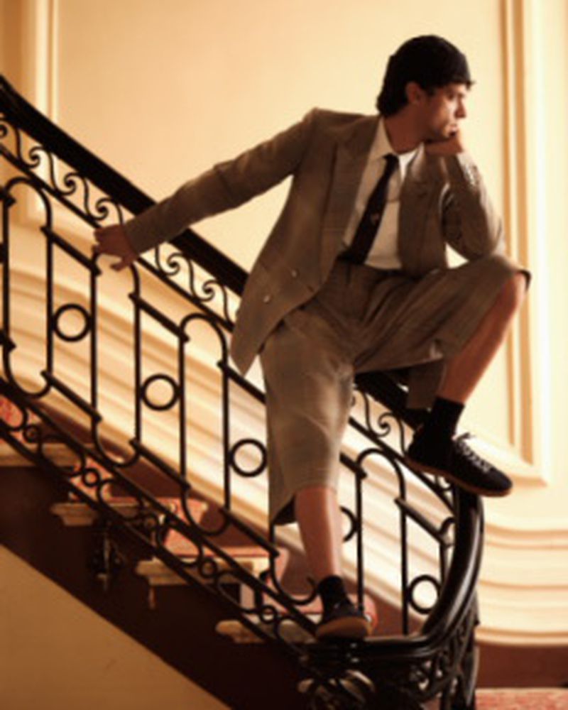 Model Marin Fabre posing on an ornate black wrought iron staircase railing wearing a tailored beige suit with wide shorts and a beanie.