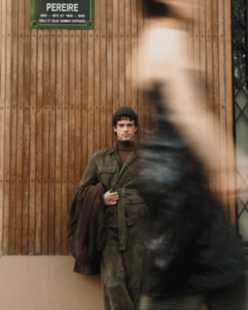 Model Marin Fabre stands against a vertical wood slat wall wearing an olive belted jacket, with a blurred figure passing in foreground.