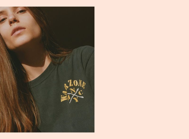 Close-up portrait of a woman in a dark green graphic t-shirt looking up in sharp direct sunlight against a pale pink border.