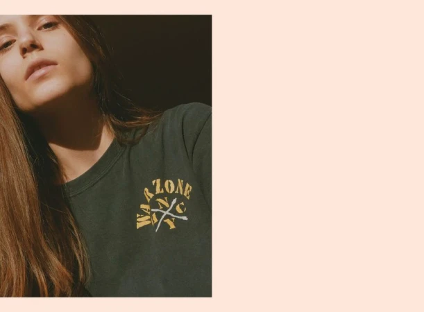 Close-up portrait of a woman in a dark green graphic t-shirt looking up in sharp direct sunlight against a pale pink border.