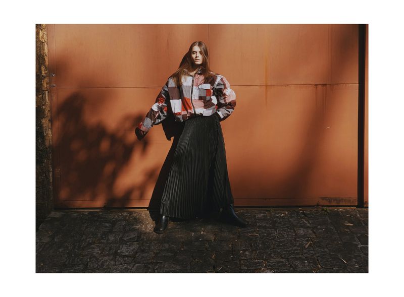 Fashion model wearing a patchwork zip jacket and black pleated maxi skirt standing on cobblestone before an orange wall.