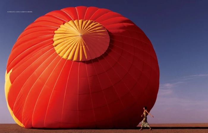 Model Qun Ye walks beside an immense red and yellow hot air balloon envelope inflated across a flat desert plain under a blue sky.